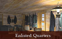 The images in this gallery provide a panoramic view of the enlisted quarters. Narrow beds line both walls. A long table with chairs runs down the center of the space. Rifles are visible in a rack and clothing is hung on hooks along the walls. The room is lit by sunlight and kerosene lamps. 