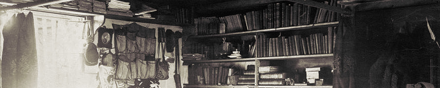 A historic photograph showing the crowded but neatly organized living area of Lt. Adolphus Greely. Books, clothing, and scientific instruments hang from walls and bookcases.