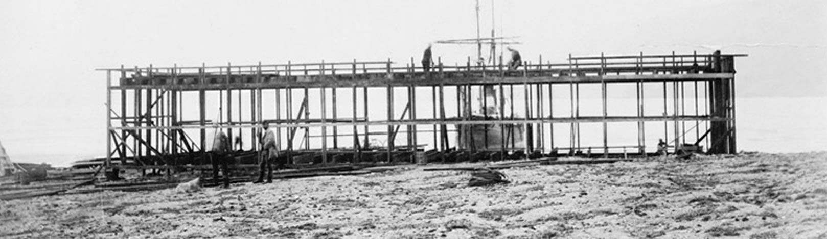 Historic image of Lady Franklin Bay Expedition House under construction. The bow of the Proteus is visible in Discovery Harbor. Two figures stand on the wooden frame of the building. One figure is visible near the sill plate.