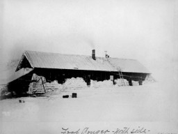A historic photograph of the Lady Franklin Bay Expedition House, with snow banked around the walls of dwelling, and a ladder leading up to the roof. 