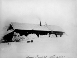 A historic photograph showing the Lady Franklin Bay Expedition house in winter. The long single store structure has snow  banked against the walls, and a ladder leads to the roof. 