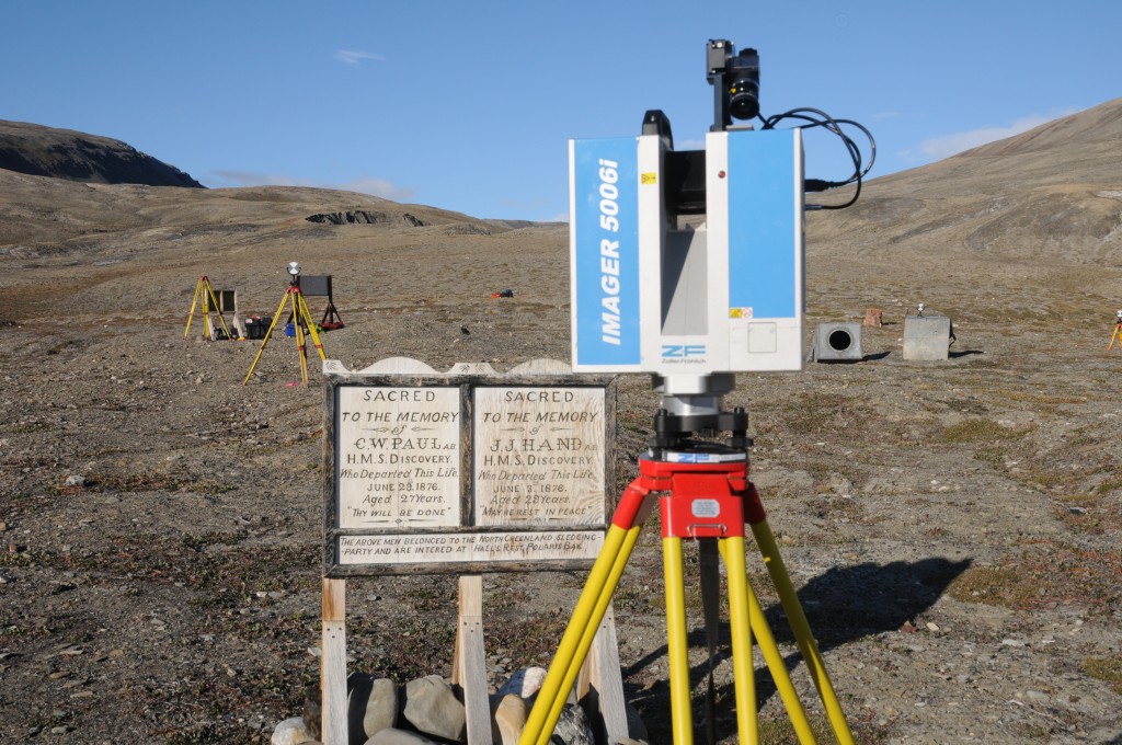 Laser Scanning: A photograph showing the laser scanner sitting atop its tripod. The scanner is rectangular in shape. In the distance are two tripods with circular target. 